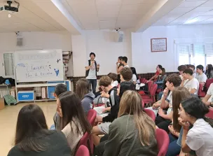 Grupo de estudiantes asistiendo a una charla o presentación en una sala, mientras un joven expone junto a una pizarra con un mensaje de bienvenida.