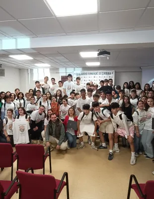 Grupo grande de estudiantes y docentes posando juntos en una sala de actos o aula de conferencias. Varias personas llevan camisetas blancas con un di