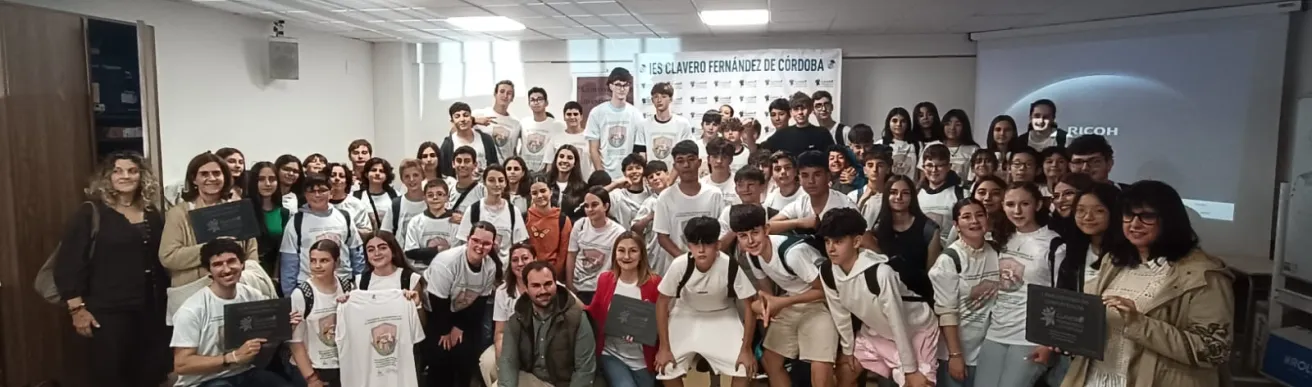 Grupo grande de estudiantes y docentes posando juntos en una sala de actos o aula de conferencias. Varias personas llevan camisetas blancas con un di