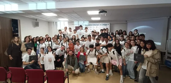 Grupo grande de estudiantes y docentes posando juntos en una sala de actos o aula de conferencias. Varias personas llevan camisetas blancas con un di