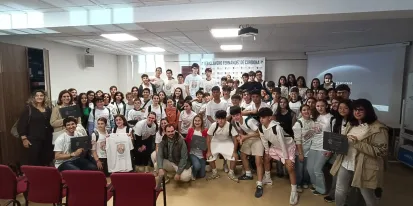 Grupo grande de estudiantes y docentes posando juntos en una sala de actos o aula de conferencias. Varias personas llevan camisetas blancas con un di